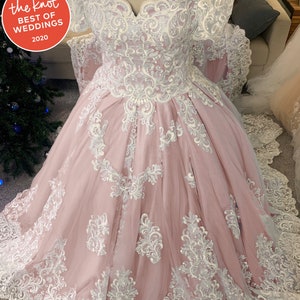 May include: A blush pink wedding dress with detailed white lace. The off-the-shoulder bodice and full skirt are decorated with floral and scrollwork patterns. The dress has a sweetheart neckline and a long train. The Knot Best of Weddings 2020 logo is visible.