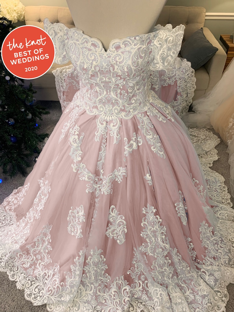 May include: A blush pink wedding dress with detailed white lace. The off-the-shoulder bodice and full skirt are decorated with floral and scrollwork patterns. The dress has a sweetheart neckline and a long train. The Knot Best of Weddings 2020 logo is visible.