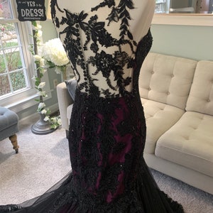 May include: A black and burgundy mermaid style gown with black lace and beading. The gown has a sheer bodice with a lace overlay and a fitted skirt.