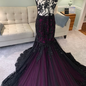 May include: A black and purple mermaid-style gown with lace detailing and a sheer bodice. The gown has a fitted silhouette and a long train.