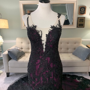 May include: A black and purple mermaid-style gown with black lace detailing and a sheer bodice. The gown has a fitted silhouette and a flowing skirt.