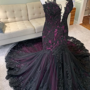 May include: A long, black and purple mermaid-style gown with black lace and beading. The gown has a sweetheart neckline and a sheer bodice.