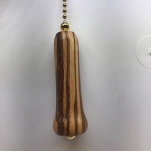 May include: A wooden fan pull with a gold chain and a gold bead at the top. The wood has a striped pattern in shades of brown and light brown.