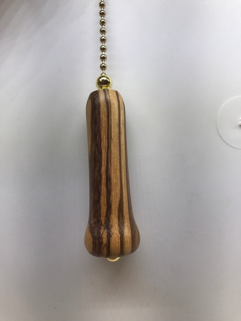K&ouml;nnte beinhalten: Ein Holz-L&uuml;fterzug mit einer goldenen Kette und einer goldenen Perle oben. Das Holz hat ein gestreiftes Muster in Braun- und Hellbraunt&ouml;nen.