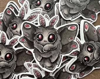 May include: A collection of stickers featuring a cartoon moth with large red eyes and pink accents on its wings and antennae. The moth is primarily gray with white outlines, creating a whimsical and artistic design. The stickers are scattered on a wooden surface.