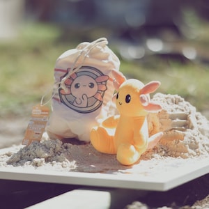 May include: A yellow axolotl plush toy with pink gills sits on a white surface with a white drawstring bag with a pink axolotl illustration and the text "Youth" printed on it. The bag and toy are surrounded by sand.