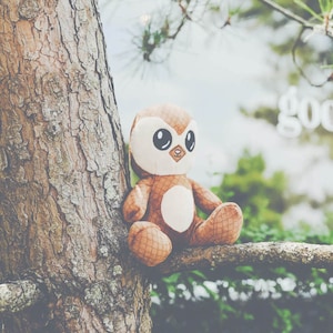 Pode incluir: Um brinquedo de pelúcia coruja marrom e branco com olhos grandes e uma barriga redonda sentado em um galho de árvore.
