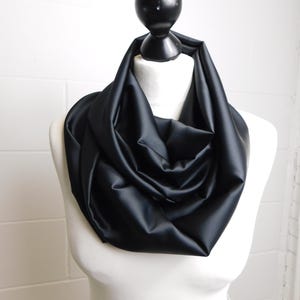 May include: A solid black infinity scarf draped in loose, voluminous folds around the neck. The fabric has a smooth, semi-matte finish with a consistent dark tone throughout, creating a structured yet soft appearance around the collar area.