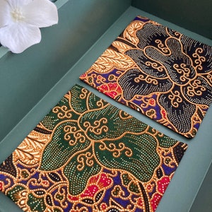 May include: Two fabric squares with a traditional Indonesian batik pattern. One square is black with gold and white accents, the other is green with gold and white accents.