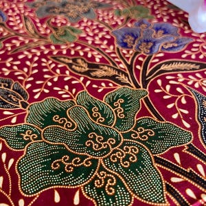 Op de afbeelding: Een close-up van een rode stof met een groen, blauw en gouden bloemenpatroon. De stof heeft een textuur en lijkt van zijde te zijn.