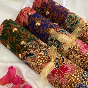 May include: Three rolls of fabric with colorful floral patterns, tied together with a gold ribbon. The fabrics are green and brown, purple and brown, and red and brown.