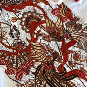 Op de afbeelding: Witte stof met een bruin, rood en zwart batikpatroon met een gestileerde vogel met een lange staart en bloemmotieven.