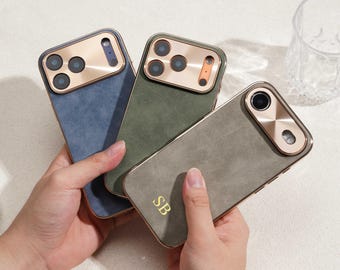 Op de afbeelding: Drie telefoonhoesjes in blauw, groen en grijs, elk met een goudkleurig camerakader en -afwerking. De grijze hoes heeft de initialen "SB" in goud. De hoesjes hebben een textuur.