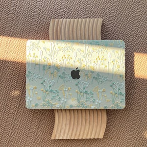May include: A laptop with a floral design cover featuring yellow, white, and teal embroidered flowers. The laptop rests on a light brown, textured wooden stand, set against a brown herringbone patterned surface.
