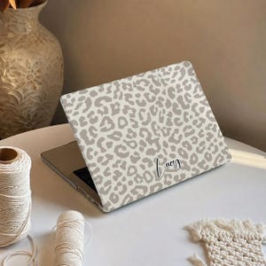 May include: A laptop skin featuring a light grey and white leopard print pattern with the name Lucy written in a black cursive script font. The skin covers the outer lid of a closed laptop, displaying a repeating animal spot design.