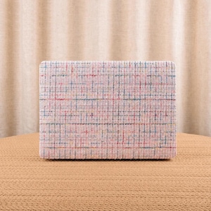 May include: A rectangular box covered in a light pink tweed fabric featuring a woven grid pattern of thin blue, yellow, and red threads. The box has a structured shape with sharp corners and a textured surface created by the interlaced multicolored yarn.