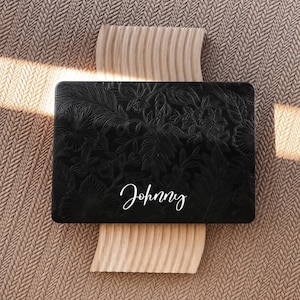 Op de afbeelding: Een rechthoekige zwarte laptophoes met een reliëf bloemenpatroon over het oppervlak. De naam Johnny is in witte schrijfletters in het midden onderaan de hoes gedrukt. Het ontwerp toont gedetailleerde blad- en bloemmotieven.