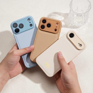 Może przedstawiać: Trzy skórzane etui na telefon w kolorze jasnoniebieskim, beżowym i kremowym. Każde etui ma wycięcie na aparat i teksturowane wykończenie. Kremowe etui ma złote inicjały "SB". Przezroczysty szklany kubek w tle.