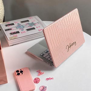 May include: A light pink laptop case with a textured crocodile skin pattern and the name Johnny written in black cursive script. The case is attached to a laptop, accompanied by a matching light pink phone case and several small stickers.