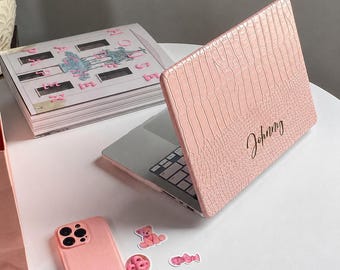 May include: A light pink laptop case with a textured crocodile skin pattern and the name Johnny written in black cursive script. The case is attached to a laptop, accompanied by a matching light pink phone case and several small stickers.
