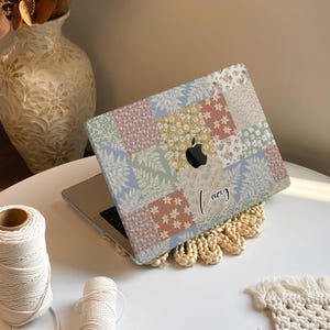 May include: A laptop with a colorful patchwork design on the lid. The design features a variety of floral patterns and colors, including pink, blue, green, and yellow. The laptop has a black Apple logo and the name "Lucy" is written in cursive on the lid.
