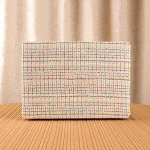 May include: A rectangular box covered in a textured, cream-coloured fabric featuring a multi-coloured grid pattern. The woven material displays thin, intersecting lines in shades of red, blue, and yellow, creating a subtle, repeating plaid design.