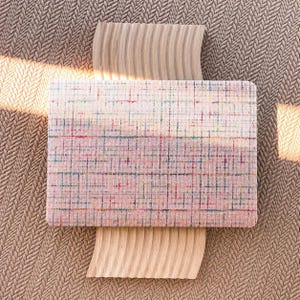 May include: A rectangular fabric panel featuring a woven plaid pattern in shades of pink, white, blue, and red. The panel is positioned over two light-colored, curved wooden pieces with a ribbed texture, all set against a brown braided surface.