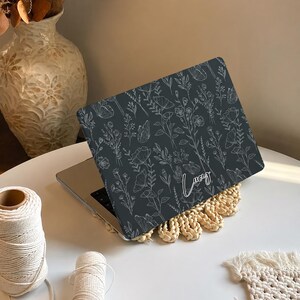 May include: A dark gray laptop skin featuring a white line-art pattern of various wildflowers, stems, and butterflies. The name Lucy is printed in a white cursive script font on the lower right side of the laptop cover, which is applied to a laptop.