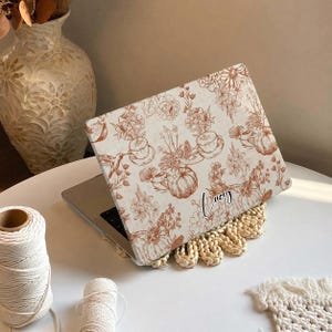 May include: A laptop skin featuring a cream background with a repeating copper-colored line art pattern of pumpkins, flowers, hummingbirds, and coffee cups. The name Lucy is printed in a black cursive script on the lower right side of the skin.