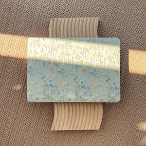 May include: A rectangular light green plaque featuring a raised, textured pattern of various wildflowers in shades of yellow, white, and sage green. The plaque is positioned on a light brown, wavy, vertical wooden base with a ribbed surface.