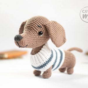 May include: A brown crocheted dachshund wearing a white and blue striped jumper. The dachshund is standing on a white surface.