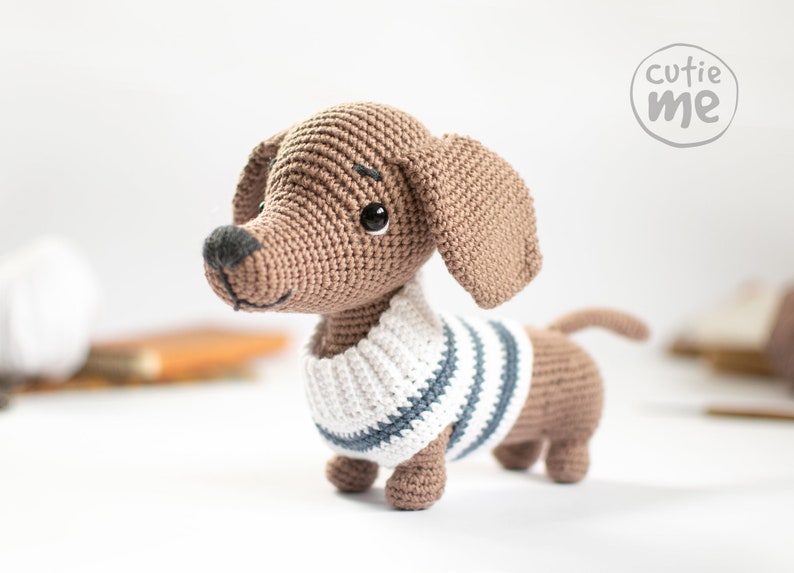 May include: A brown crocheted dachshund wearing a white and blue striped jumper. The dachshund is standing on a white surface.