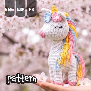 May include: A crocheted unicorn toy with a white body, pink nose and hooves, and a colorful mane and tail. The unicorn has a yellow horn and is adorned with blue and pink flowers. The word "pattern" is visible in the image.