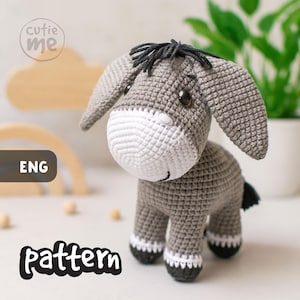 Puede incluir: Un juguete de burro tejido a ganchillo en gris, blanco y negro. El burro tiene orejas grandes, un hocico blanco y ojos negros. La palabra "pattern" está escrita en negro en la parte inferior de la imagen.