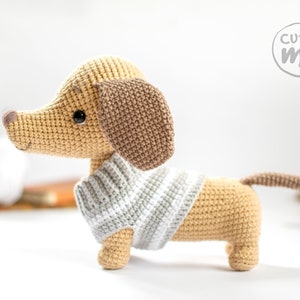 May include: A crocheted dachshund wearing a striped jumper. The dachshund is tan with brown ears and a black nose. The dachshund is standing on a white surface. The image includes the text "cutie me" in a circle.