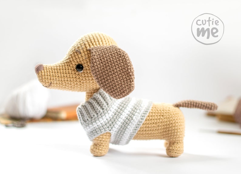 May include: A crocheted dachshund wearing a striped jumper. The dachshund is tan with brown ears and a black nose. The dachshund is standing on a white surface. The image includes the text "cutie me" in a circle.
