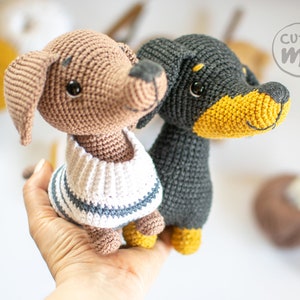 May include: Two crocheted dog plushies, one brown with a white and blue striped sweater and one black with a yellow nose. The plushies are held in a person's hand.