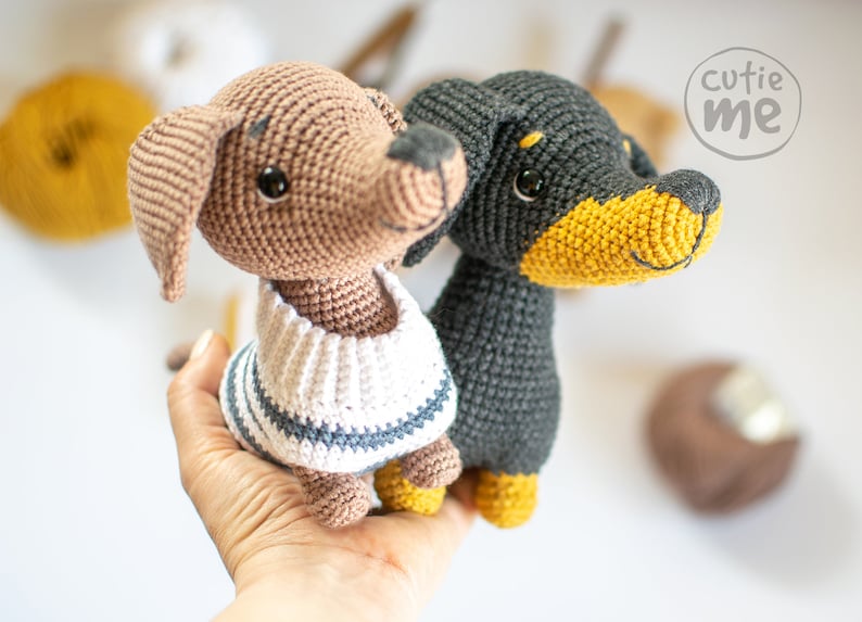 May include: Two crocheted dog plushies, one brown with a white and blue striped sweater and one black with a yellow nose. The plushies are held in a person's hand.