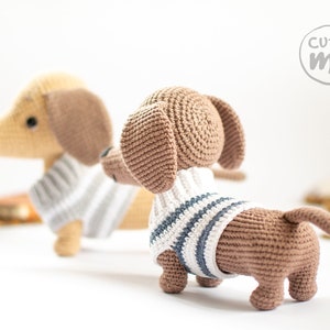 May include: Two crocheted dachshund dog plushies wearing striped sweaters. The plushies are brown and tan with black eyes and noses. The plushies are sitting on a white surface. The text "cutie me" is visible in the background.
