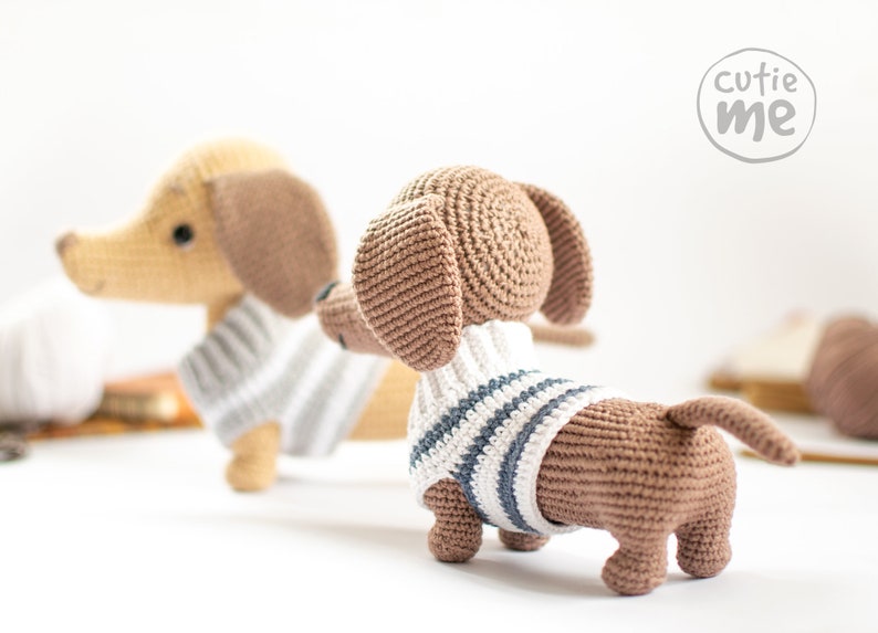 May include: Two crocheted dachshund dog plushies wearing striped sweaters. The plushies are brown and tan with black eyes and noses. The plushies are sitting on a white surface. The text "cutie me" is visible in the background.