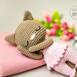 May include: A handmade crochet doll in a sleeping pose. The doll has a brown head and limbs, with a pink face and closed eyes. It wears a pink dress and rests on a pink pillow. The image includes the text "cutie me".