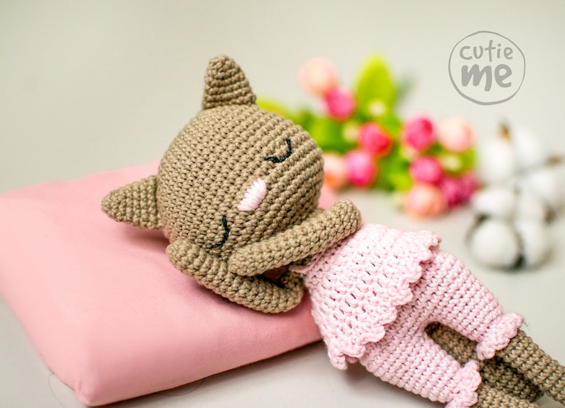 May include: A handmade crochet doll in a sleeping pose. The doll has a brown head and limbs, with a pink face and closed eyes. It wears a pink dress and rests on a pink pillow. The image includes the text "cutie me".