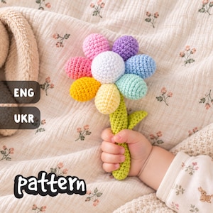 May include: A crocheted flower rattle with a white center and petals in shades of pink, purple, blue, teal, yellow, and orange. The stem is light green with a single leaf. The toy is held by a hand, next to a beige plush bunny toy.