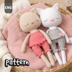 May include: Two handmade crochet cat dolls in a woven basket. One doll is beige with pink shorts, the other is white with gray overalls. Both have closed eyes and pink noses. The dolls are lying on a pink blanket.