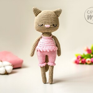 May include: A handmade crochet cat doll in a light brown color, wearing a pink outfit. The doll has closed eyes, a pink nose, and a ruffled top and pants. The doll is standing in front of a blurred background with the text "cutie me".