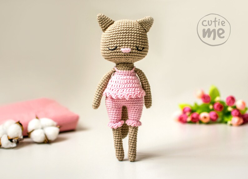May include: A handmade crochet cat doll in a light brown color, wearing a pink outfit. The doll has closed eyes, a pink nose, and a ruffled top and pants. The doll is standing in front of a blurred background with the text "cutie me".