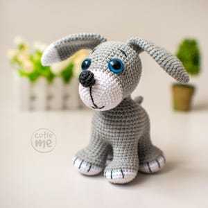 May include: A handmade crochet dog in gray, white, and black. The dog has large blue eyes, a black nose, and long floppy ears. The dog is sitting with its paws forward. The logo "cutie me" is in the lower left corner.
