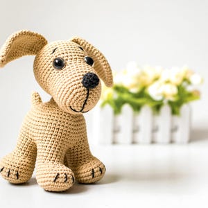 May include: A crocheted dog toy in tan yarn with black accents. The dog has large black eyes, a black nose, and a stitched smile. The ears are floppy, and the paws have black stitched details. A white picket fence with yellow flowers is in the background.