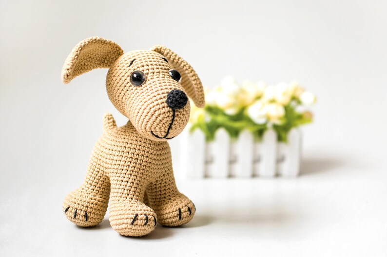 May include: A crocheted dog toy in tan yarn with black accents. The dog has large black eyes, a black nose, and a stitched smile. The ears are floppy, and the paws have black stitched details. A white picket fence with yellow flowers is in the background.