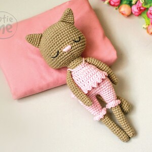 May include: A crocheted cat doll wearing pink pajamas, resting on a pink pillow. The doll has closed eyes, a pink nose, and is lying on its side. The image includes the text "cutie me" in a circle.